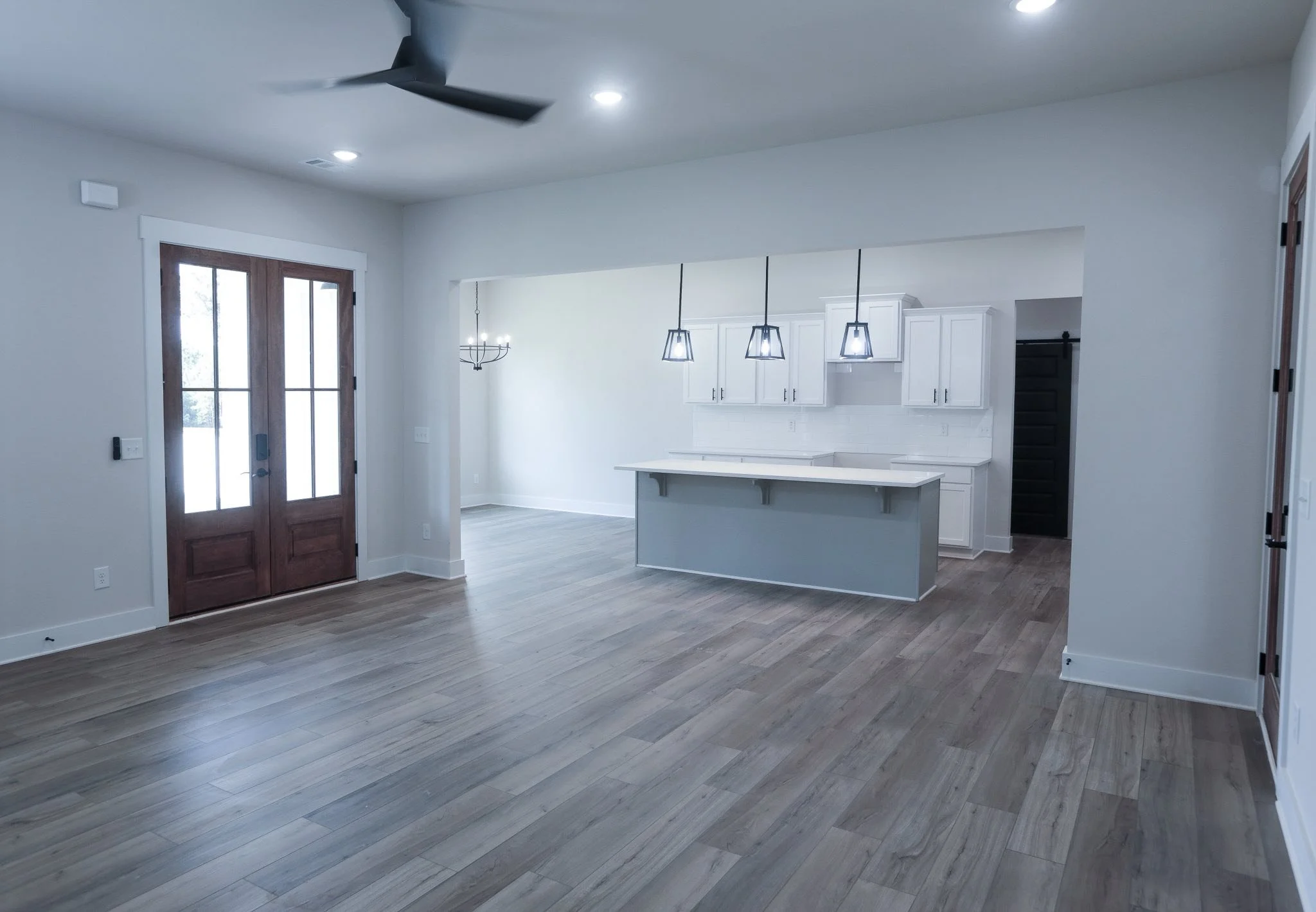 Empty modern kitchen and dining area with light wooden flooring, white cabinets, and a kitchen island, illuminated by hanging pendant lights, with a door leading outside.