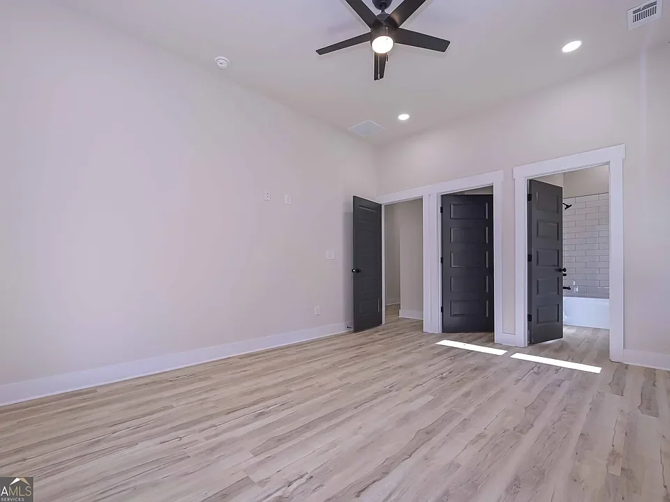 Empty bedroom with light wood flooring, white walls, and a ceiling fan. Three doorways lead to a closet, bathroom, and another room.