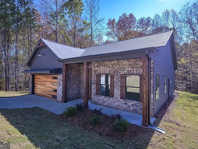 A modern house with brick and gray siding exterior, wooden accents, and a two-car garage with a wooden door, surrounded by a grassy yard and trees in the background.