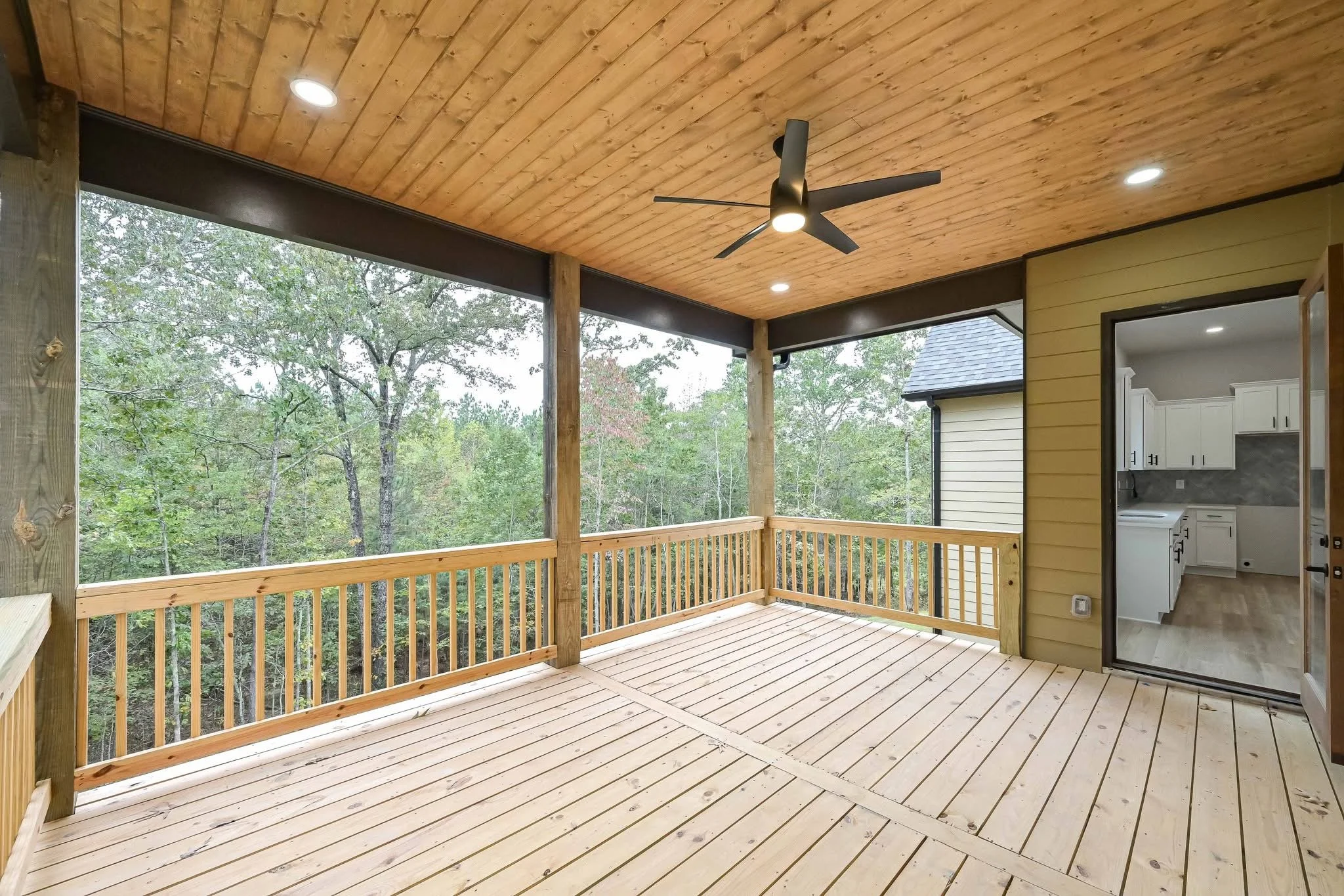 A covered wooden balcony with ceiling fan, recessed lighting, and a view of trees. Doorway leads to a kitchen with white cabinets.