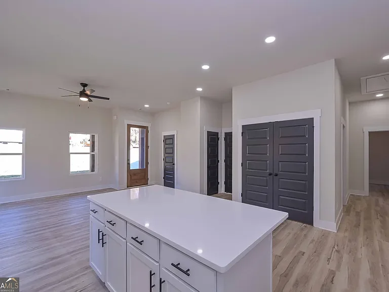 Modern kitchen with white island, light-colored wood flooring, white walls, ceiling fan, and multiple windows, with dark gray closet doors.