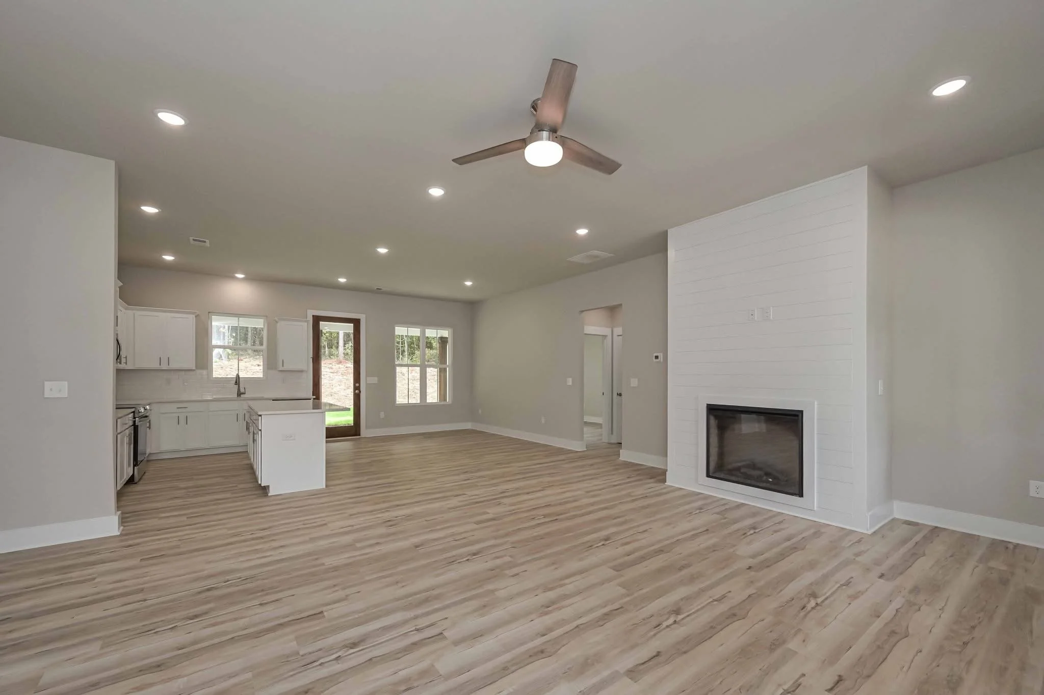 Empty living room with light-colored wood floors, white walls, a ceiling fan, built-in fireplace, and large windows, leading into a kitchen area with white cabinets.