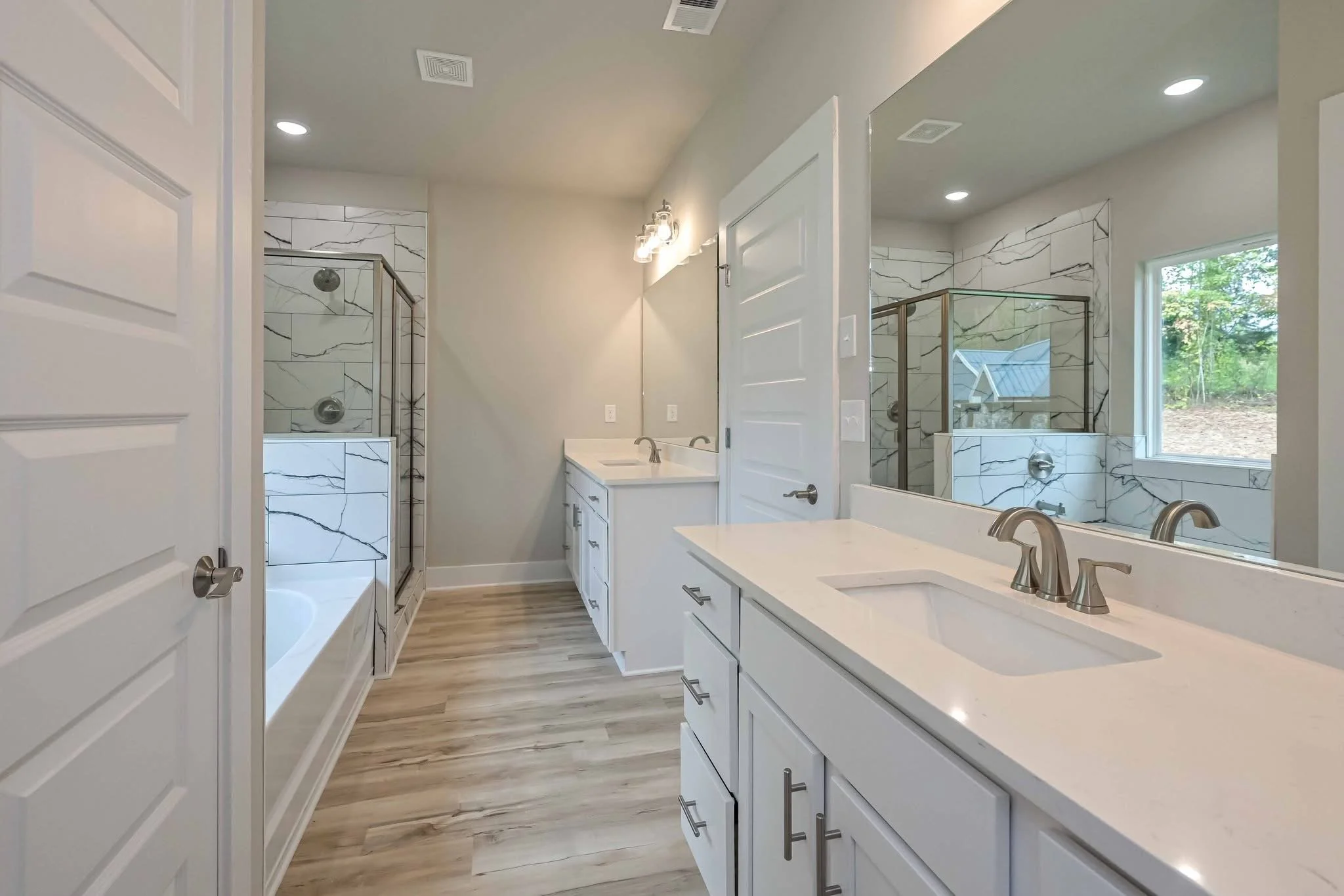 Modern bathroom with white vanity, marble shower, bathtub, and large mirror, hardwood floor, and natural light from window.