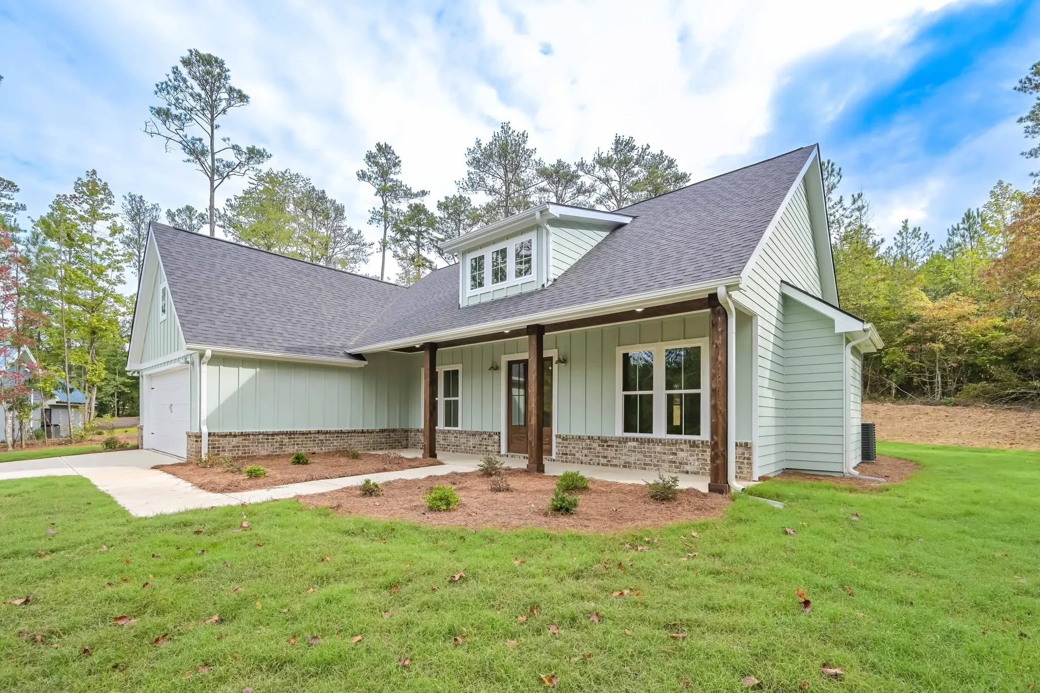 A newly constructed house with light blue exterior siding, a brick foundation, and a gabled roof. The house features a small porch with wooden posts, a front door, and multiple windows. The yard has green grass and small shrubs, set against a backdro