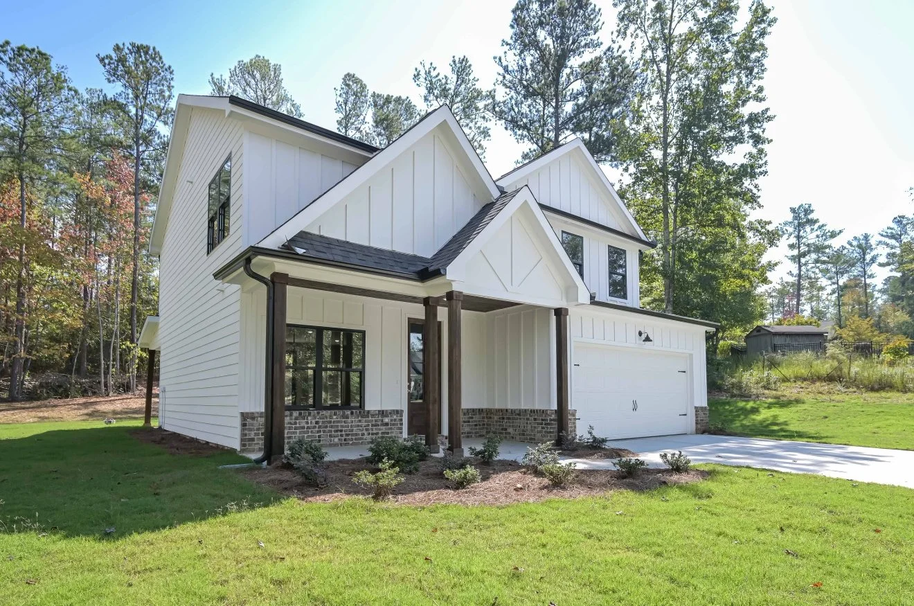 New two-story white house with black window frames, a two-car garage, and a small front porch with wooden posts, surrounded by a green lawn and trees.