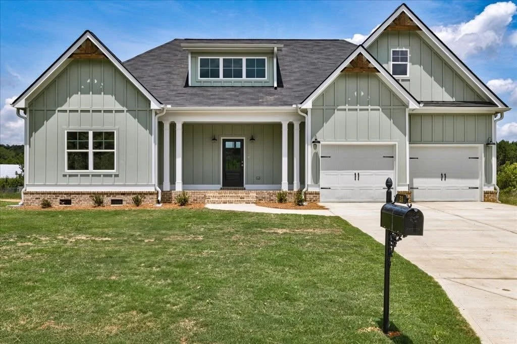 A new two-story house with a light green exterior, gray roof, and attached double garage. The front yard has a green lawn and a black mailbox on a post near the driveway.