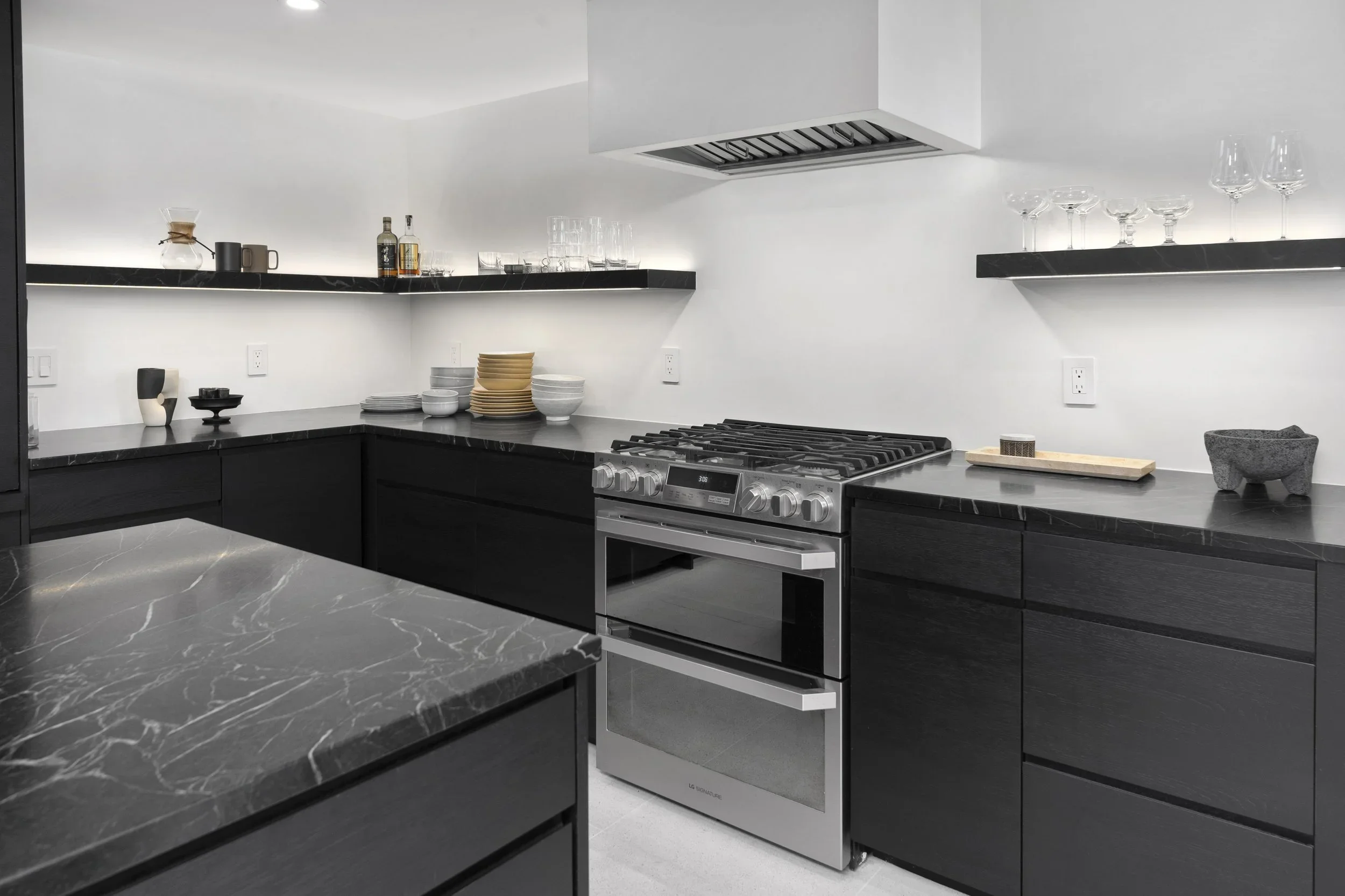 Modern kitchen with black cabinets, black marble countertops, a stainless steel stove, floating black shelves with glassware and dishes, and minimal decorative items.