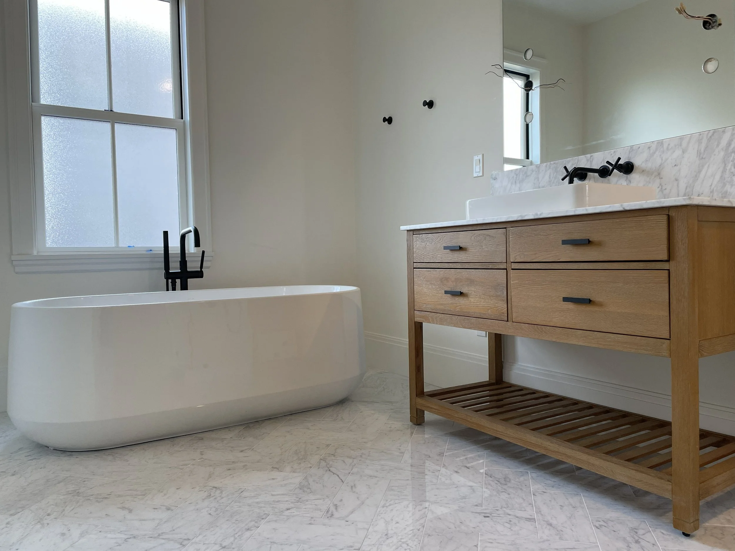Modern bathroom with a white freestanding bathtub next to a window, wooden vanity with a marble countertop, and black fixtures.