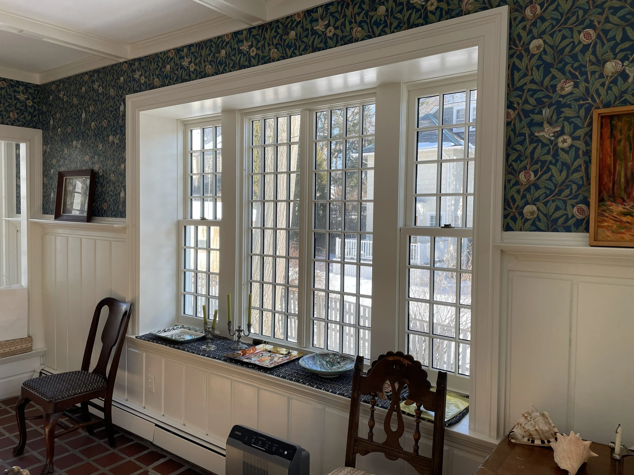 A cozy living room featuring a large bay window with multiple glass panes, decorated with wall-mounted paintings, a wooden chair, a windowsill with decorative items like candles and trays, a section of brick flooring, and a heating vent below the window.