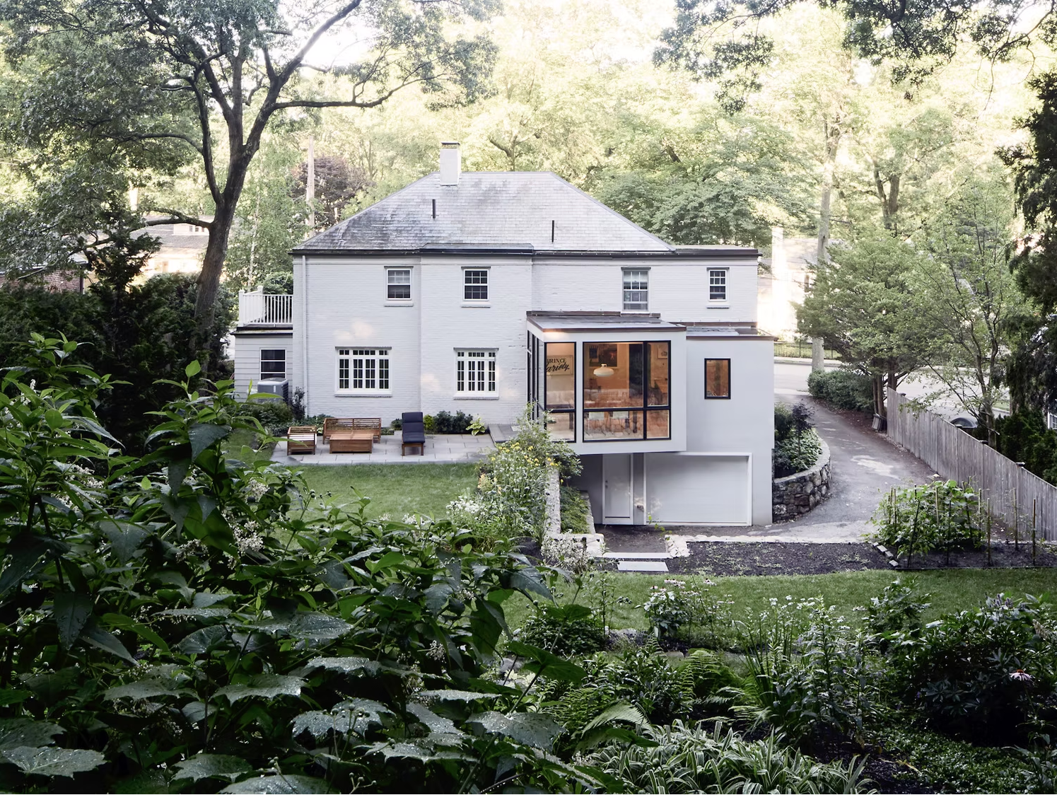 Modern white house with large windows and a backyard garden with outdoor seating, surrounded by trees, with a driveway on the side and a fenced yard.