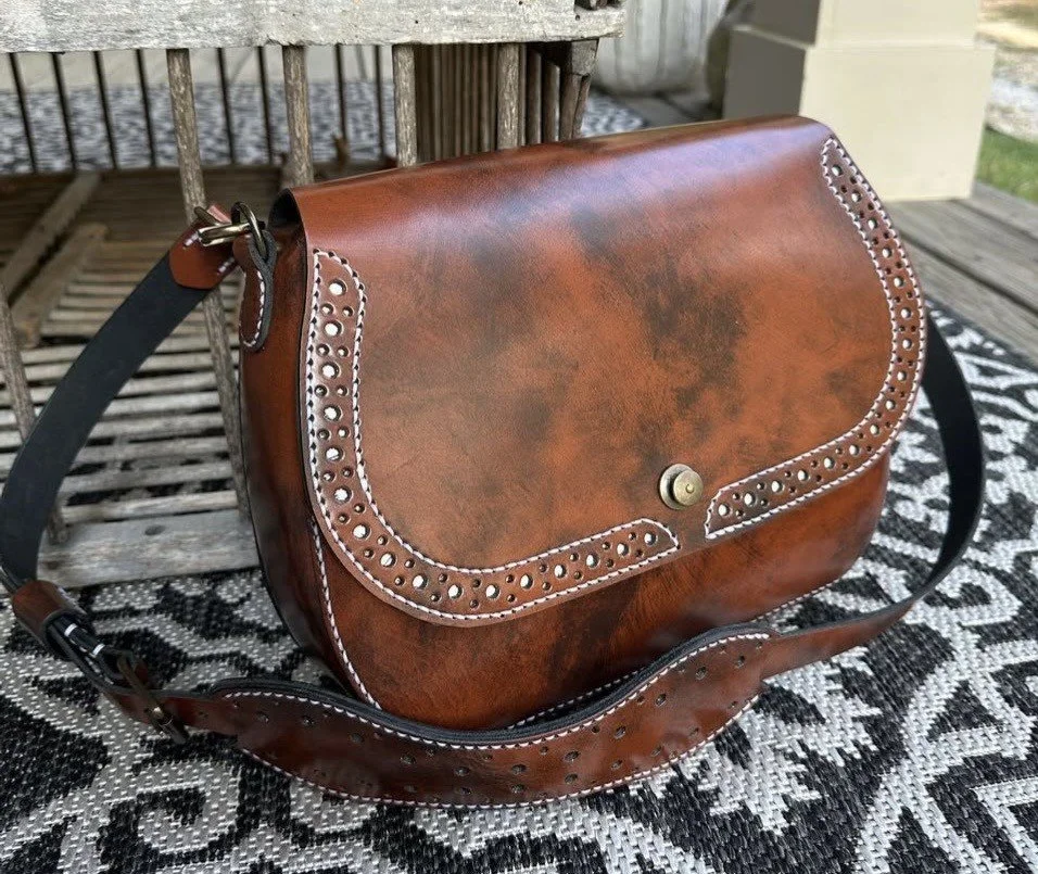 Brown leather crossbody handbag with decorative perforated trim and adjustable strap on a patterned outdoor chair