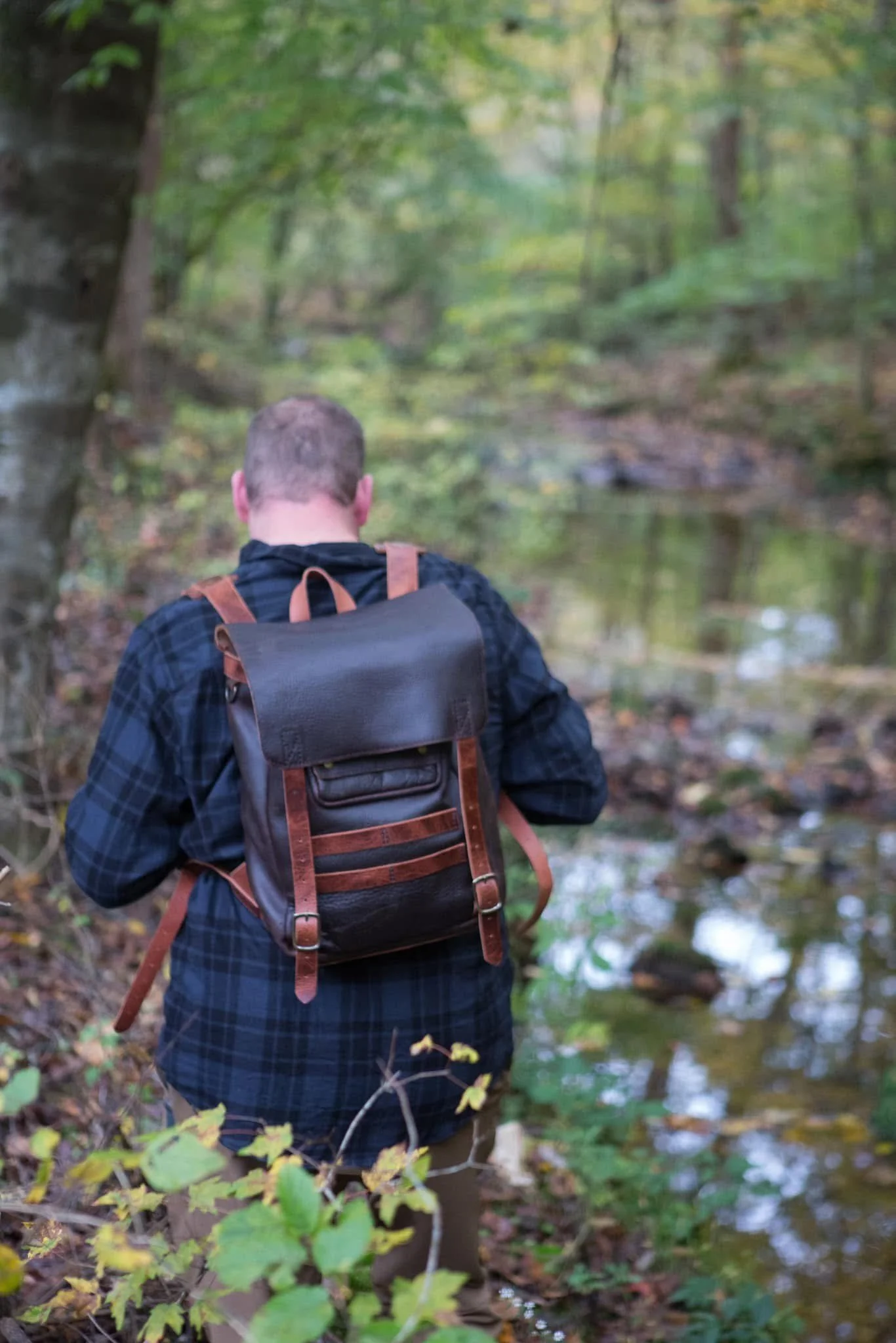 Person with a leather backpack walking beside a forest stream
