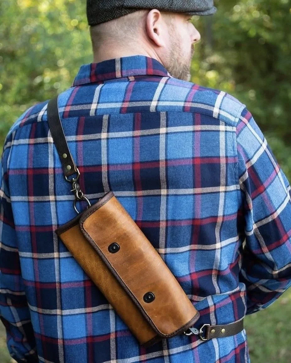 Man wearing a blue plaid shirt and flat cap, carrying a leather shoulder bag outdoors