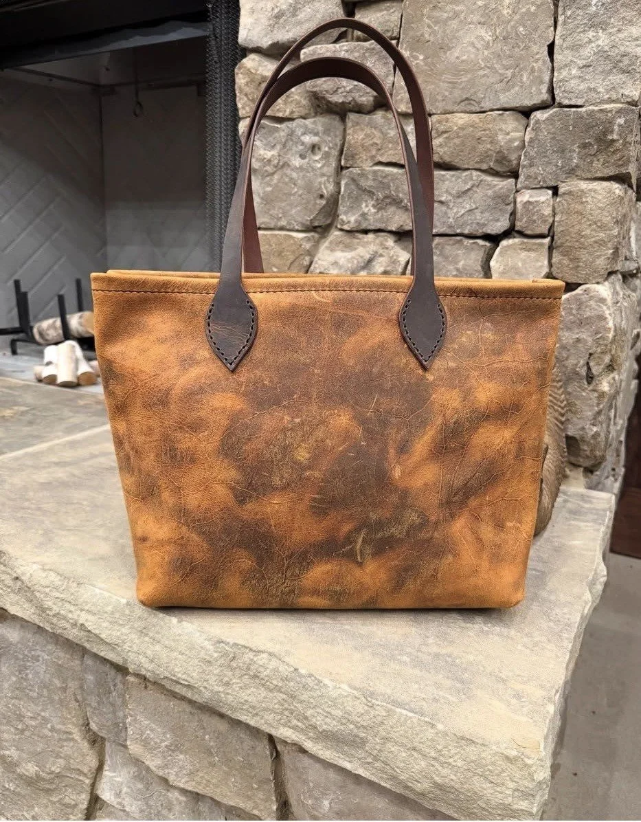 Distressed brown leather tote bag with dark leather handles displayed on a stone fireplace mantel.