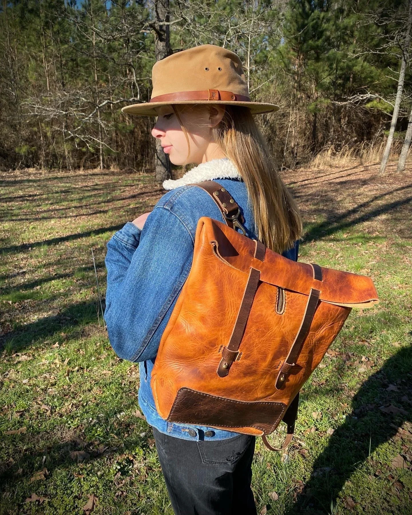 Woman wearing a wide-brimmed hat and denim jacket with a large leather backpack in a sunlit pine forest