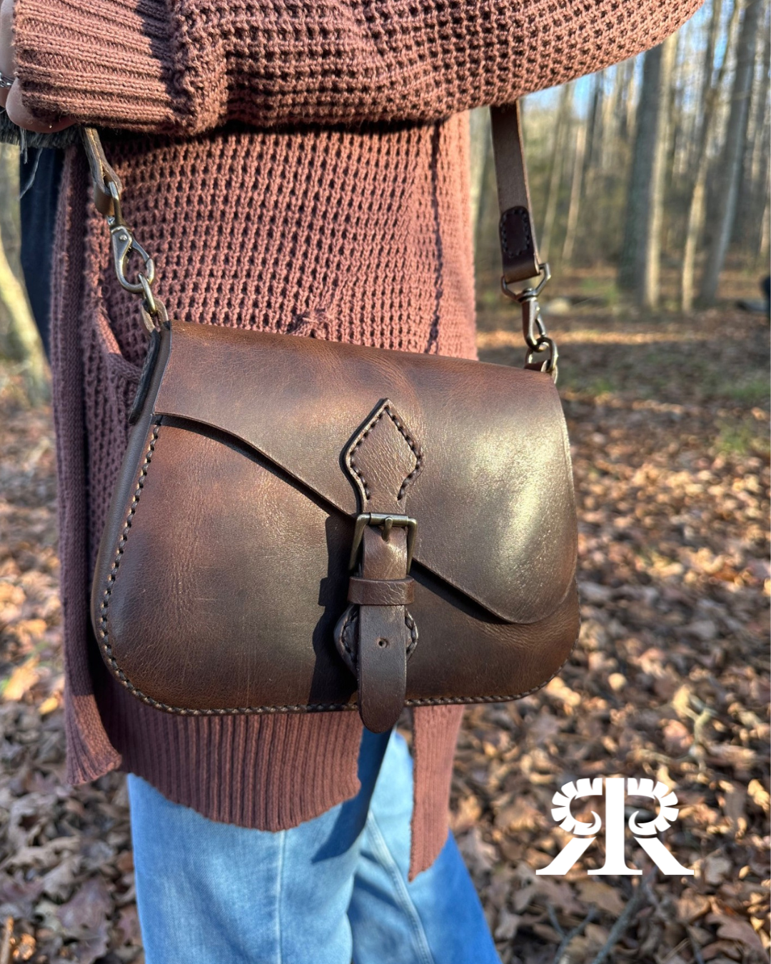 Brown leather crossbody purse with buckle closure worn over a knitted sweater in a wooded outdoor setting