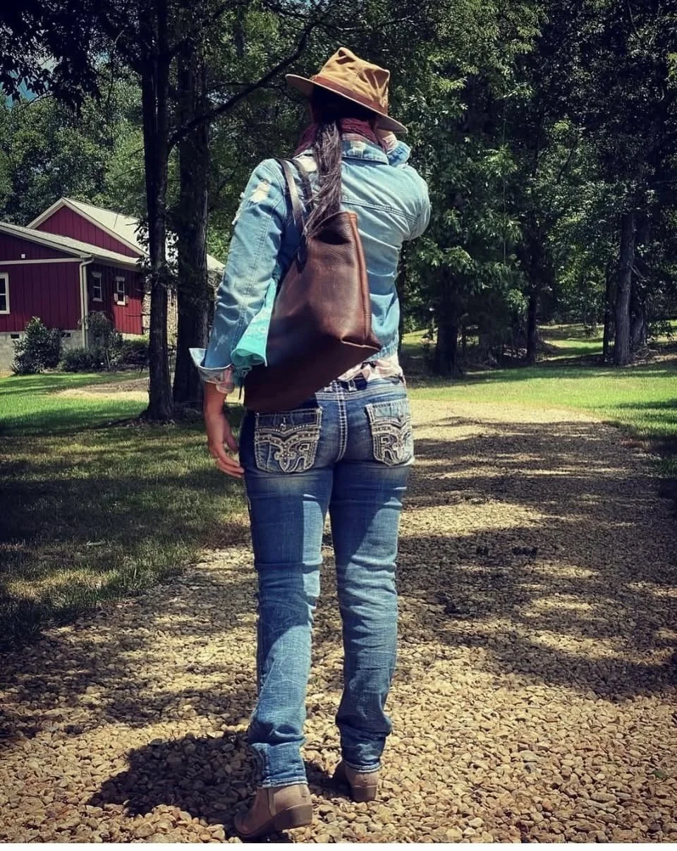 Woman wearing a hat, denim shirt, jeans, and leather backpack walking along a gravel path near a red cabin