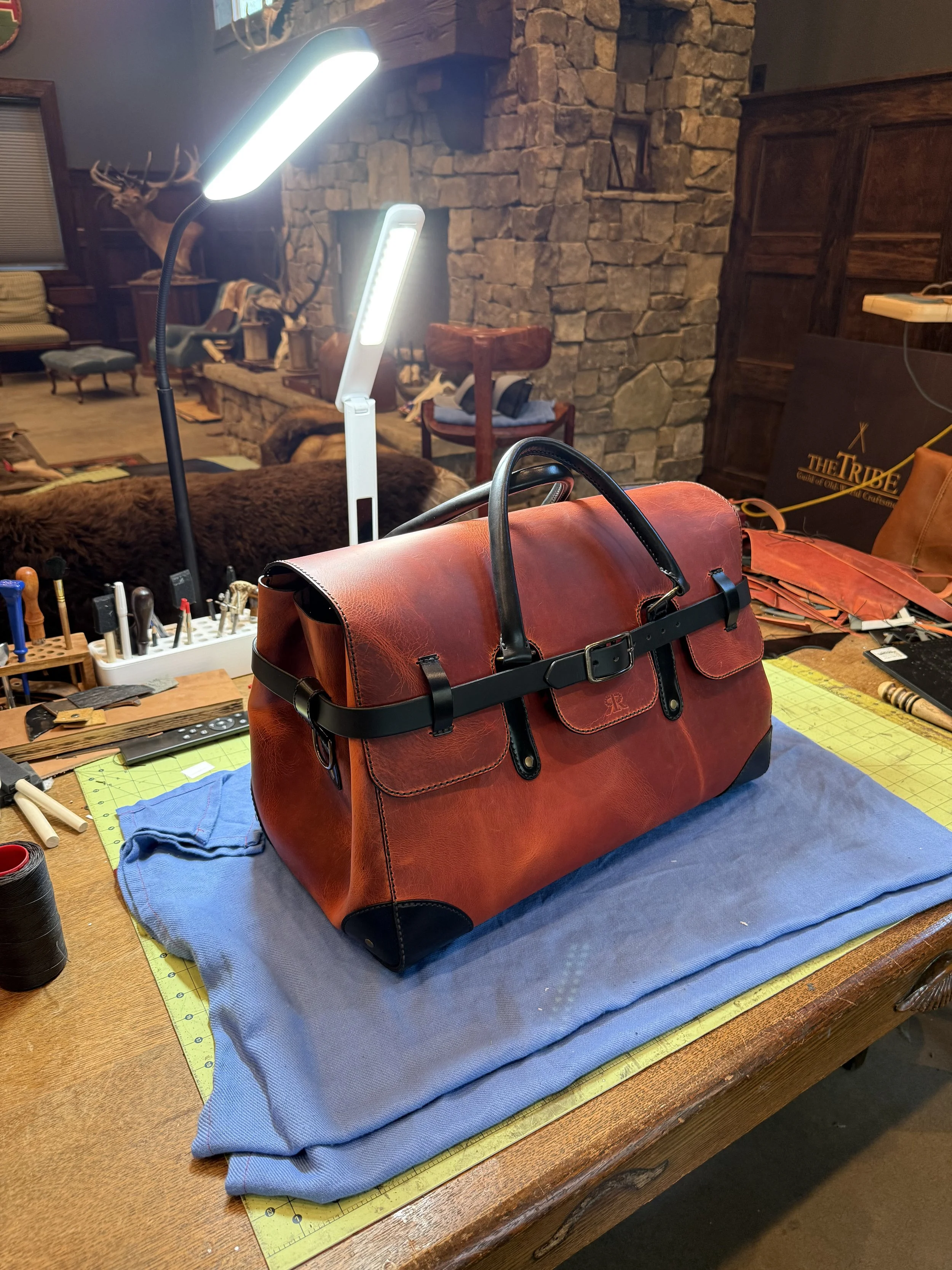 Large burgundy leather duffel bag with black straps on a workbench in a leather workshop, surrounded by tools and sewing materials. luxury bag. louis vuitton. Hermes