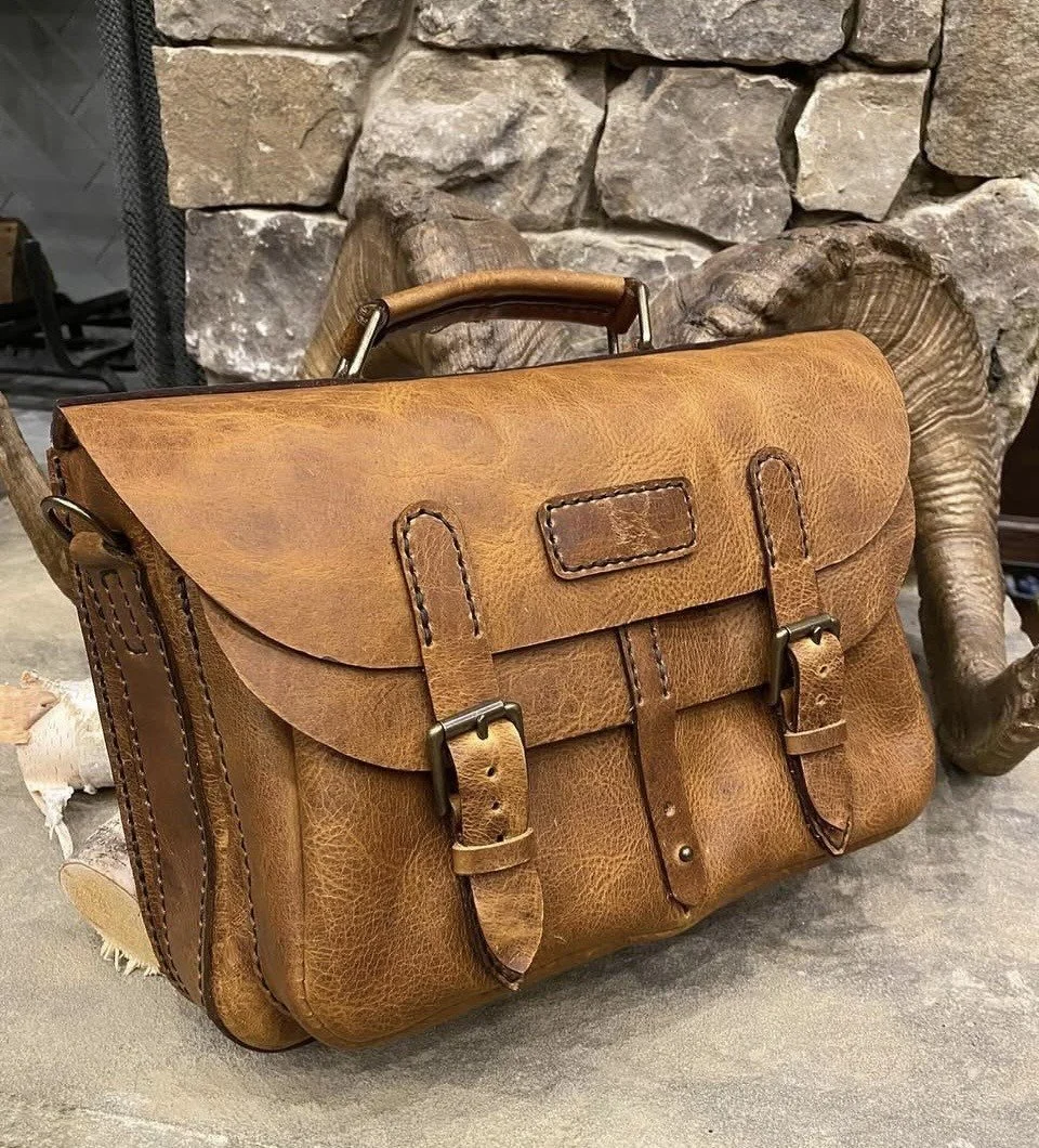 Vintage brown leather satchel bag with buckles and top handle in front of a stone fireplace