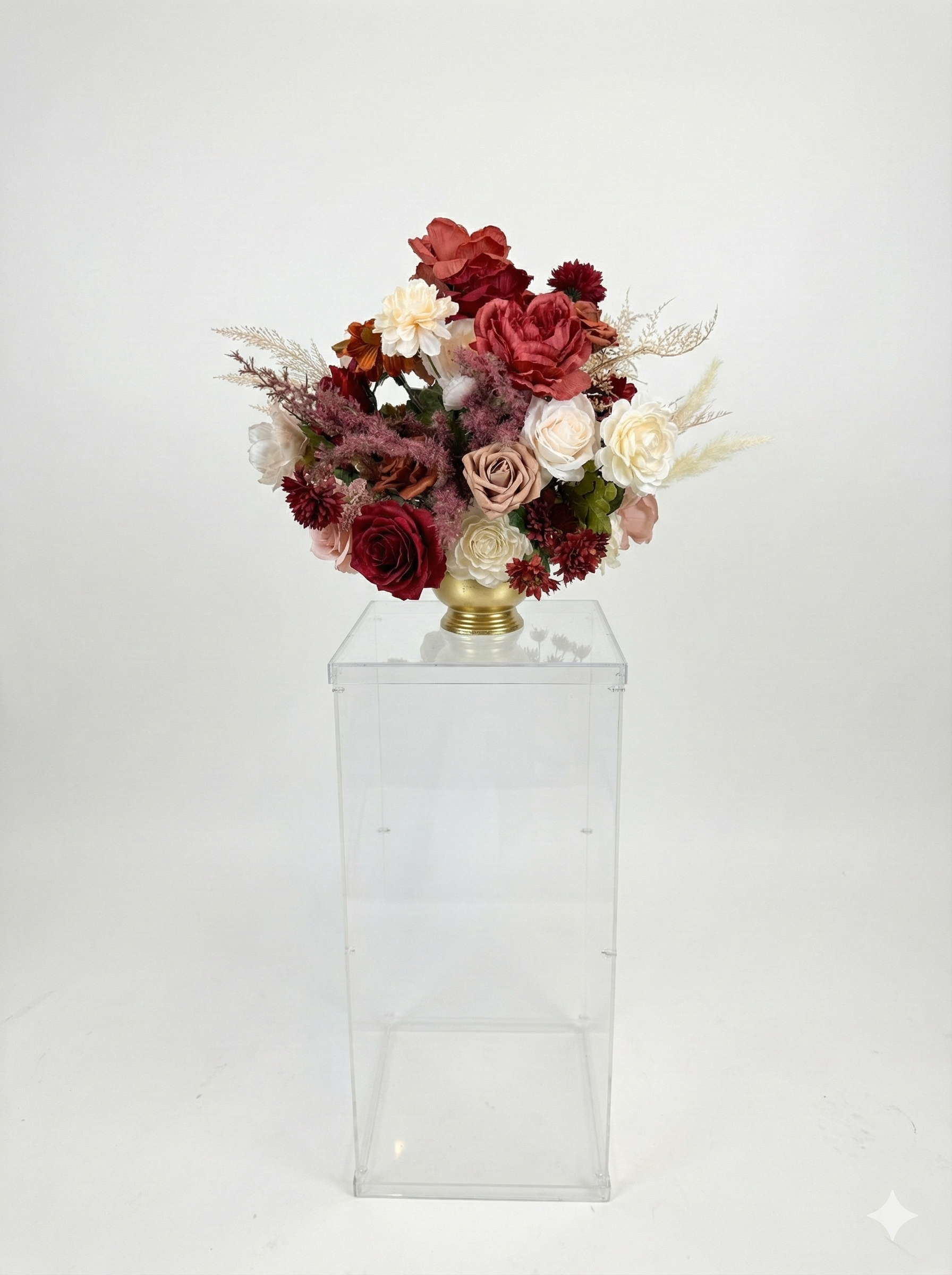 Artificial floral arrangement with roses and various flowers in a gold vase, placed on a transparent pedestal against a plain white background.