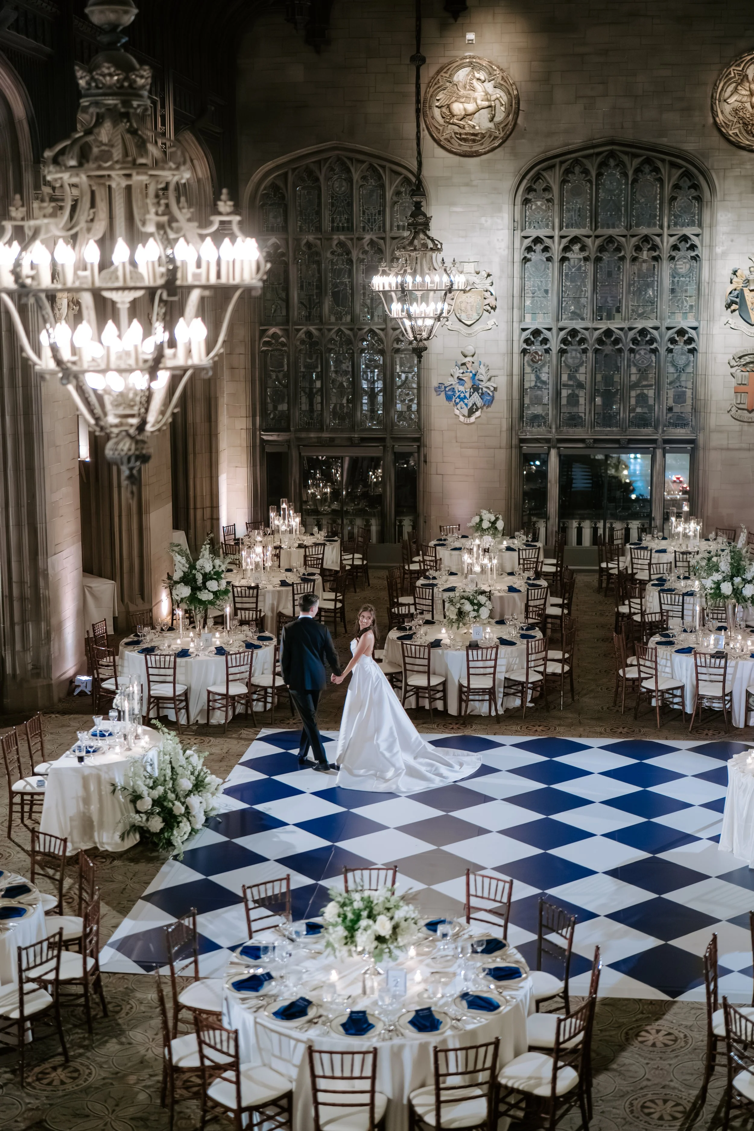A bride and groom dancing on a checkered dance floor in an elegant, historic banquet hall with high ceilings, chandeliers, and ornate wall decorations.