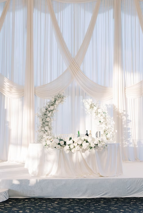 Wedding reception table with white flowers, surrounded by elegant draped white curtains and soft natural light.