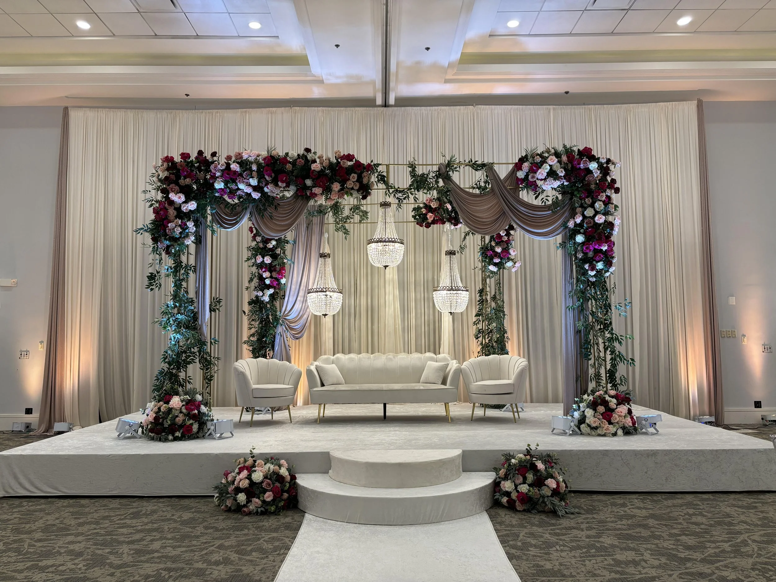 Elegant wedding stage with floral arch, chandeliers, and white furniture.