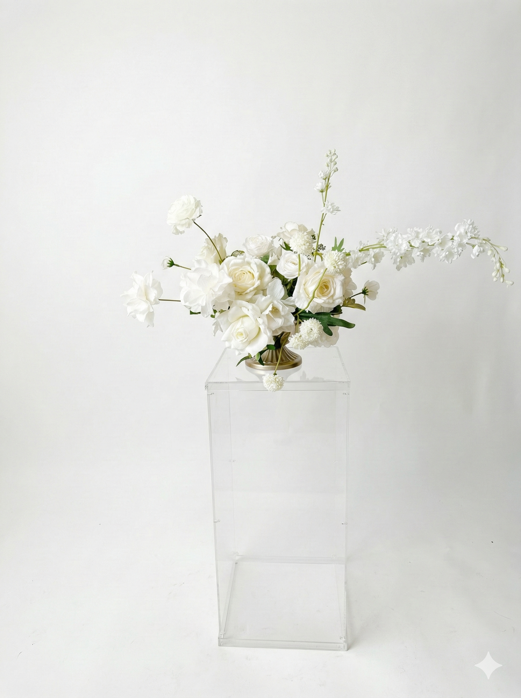 White floral arrangement with roses, orchids, and other flowers in a modern vase on a clear stand against a plain background.