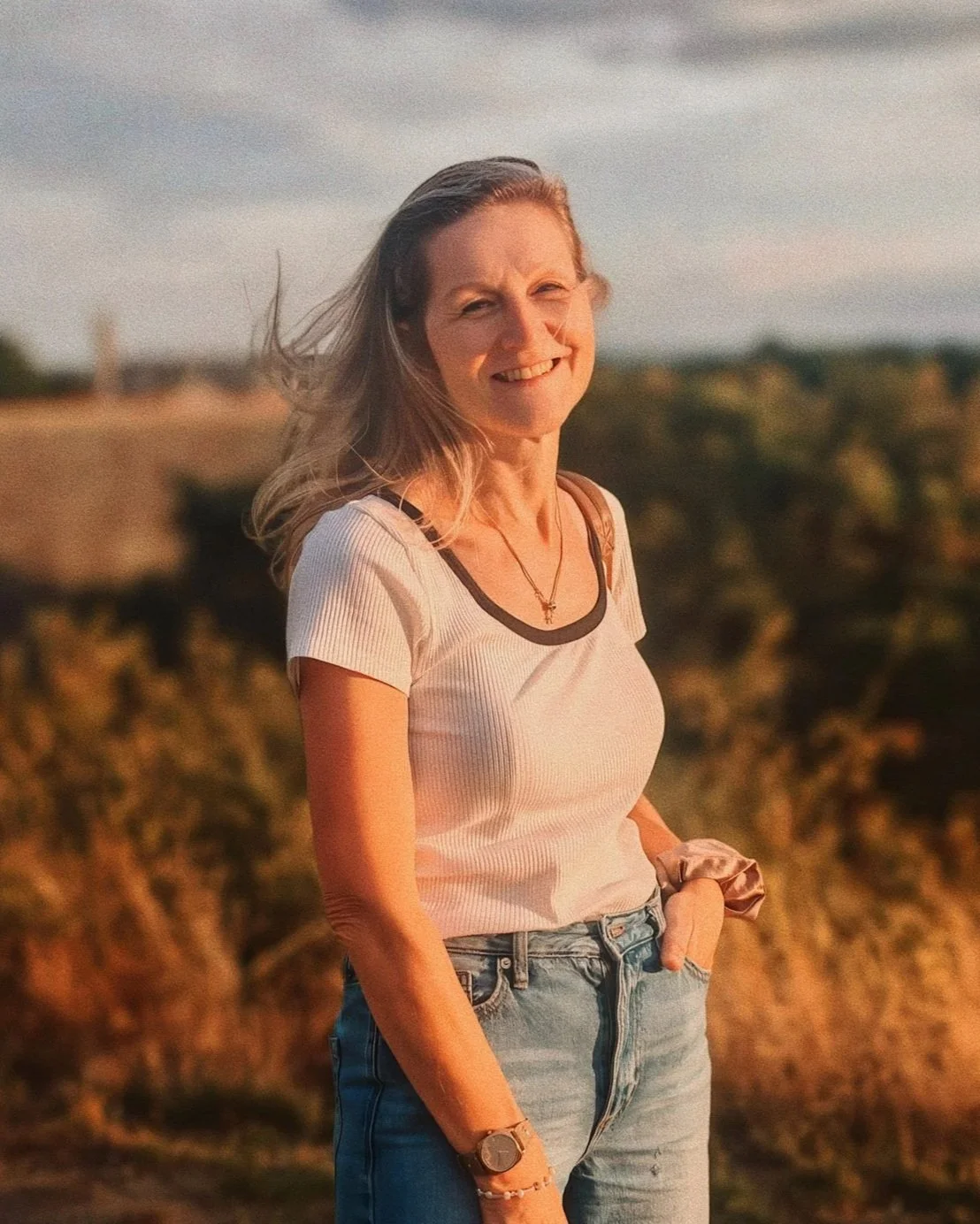 A woman standing outdoors during sunset, smiling, wearing a white short-sleeve top with black trim, blue jeans, and accessories, with a scenic blurred landscape in the background.