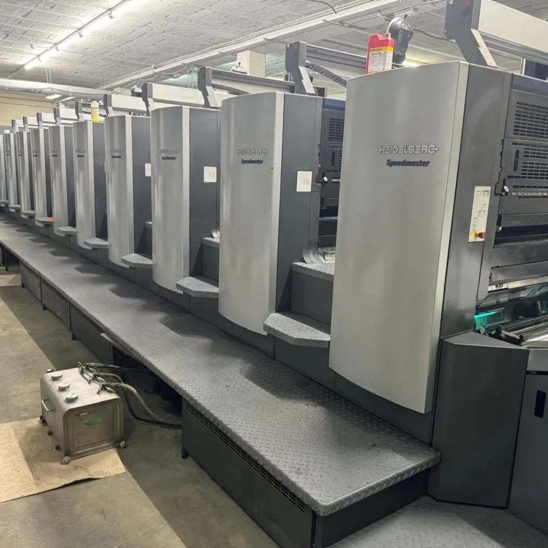 Row of Heidelberg Speedmaster printing presses in a factory or print shop, with control panels and industrial lighting overhead.