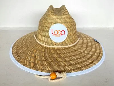 A woven straw hat with a round white sticker featuring the red Loops logo, and an orange bead on the brim.