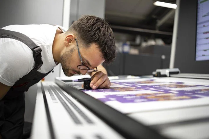 Man inspecting a printed sheet using a magnifying glass in a printing or inspection facility.