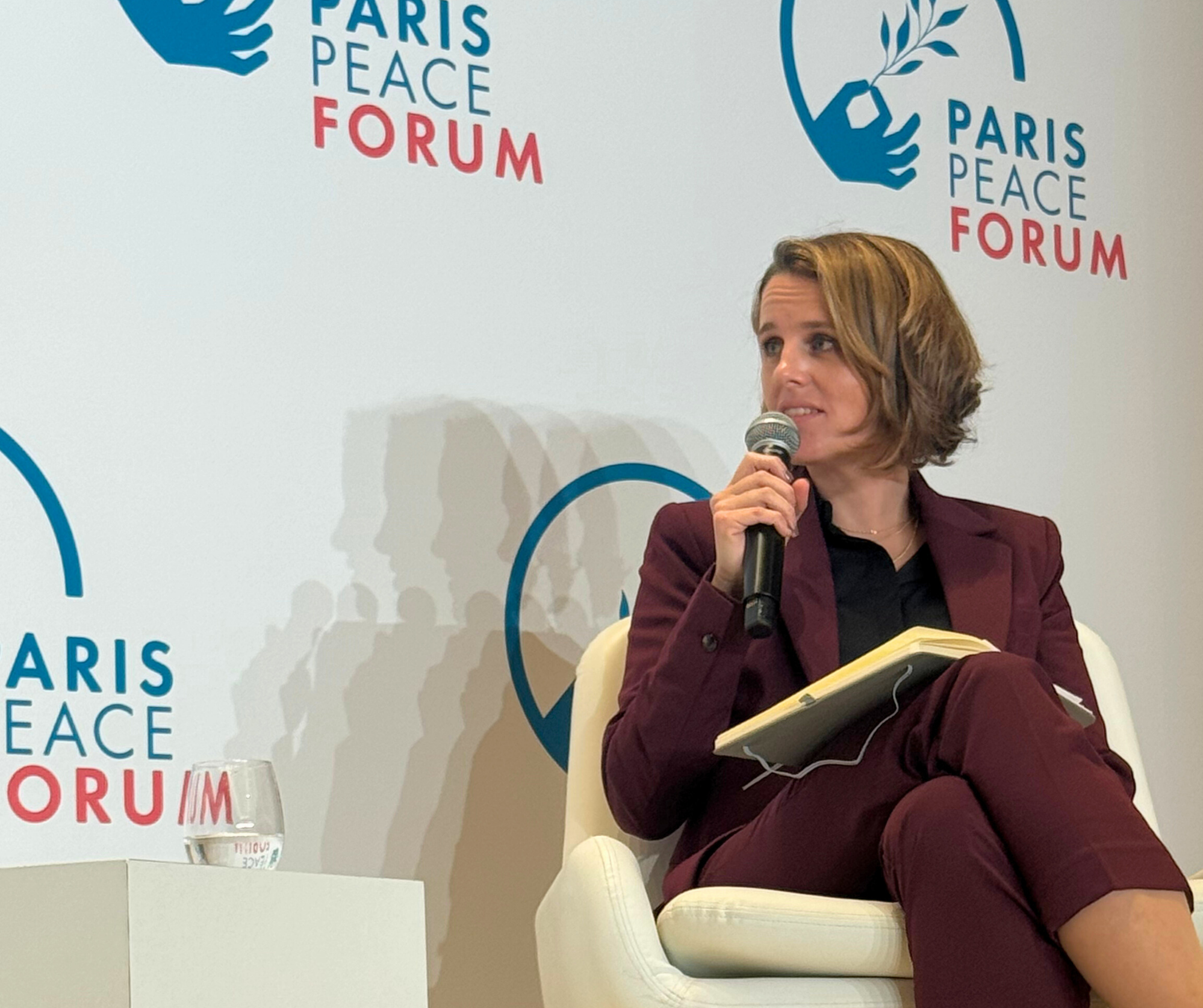 A woman with short brown hair sitting in a white chair, holding a microphone and a notebook, participating in a panel at the Paris Peace Forum. The background features a white wall with logos and text reading 'Paris Peace Forum' and a hand holding an olive branch.