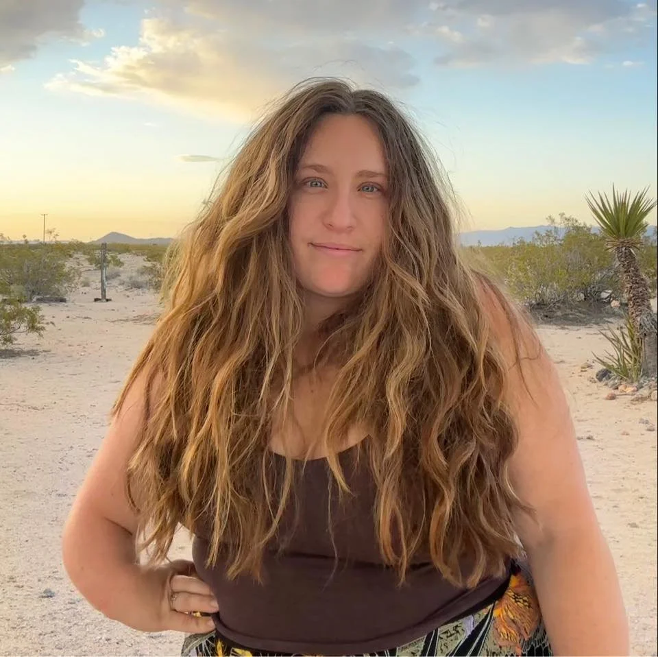 A woman with long, wavy brown hair stands in a desert landscape at sunset, wearing a black sleeveless top and patterned pants.