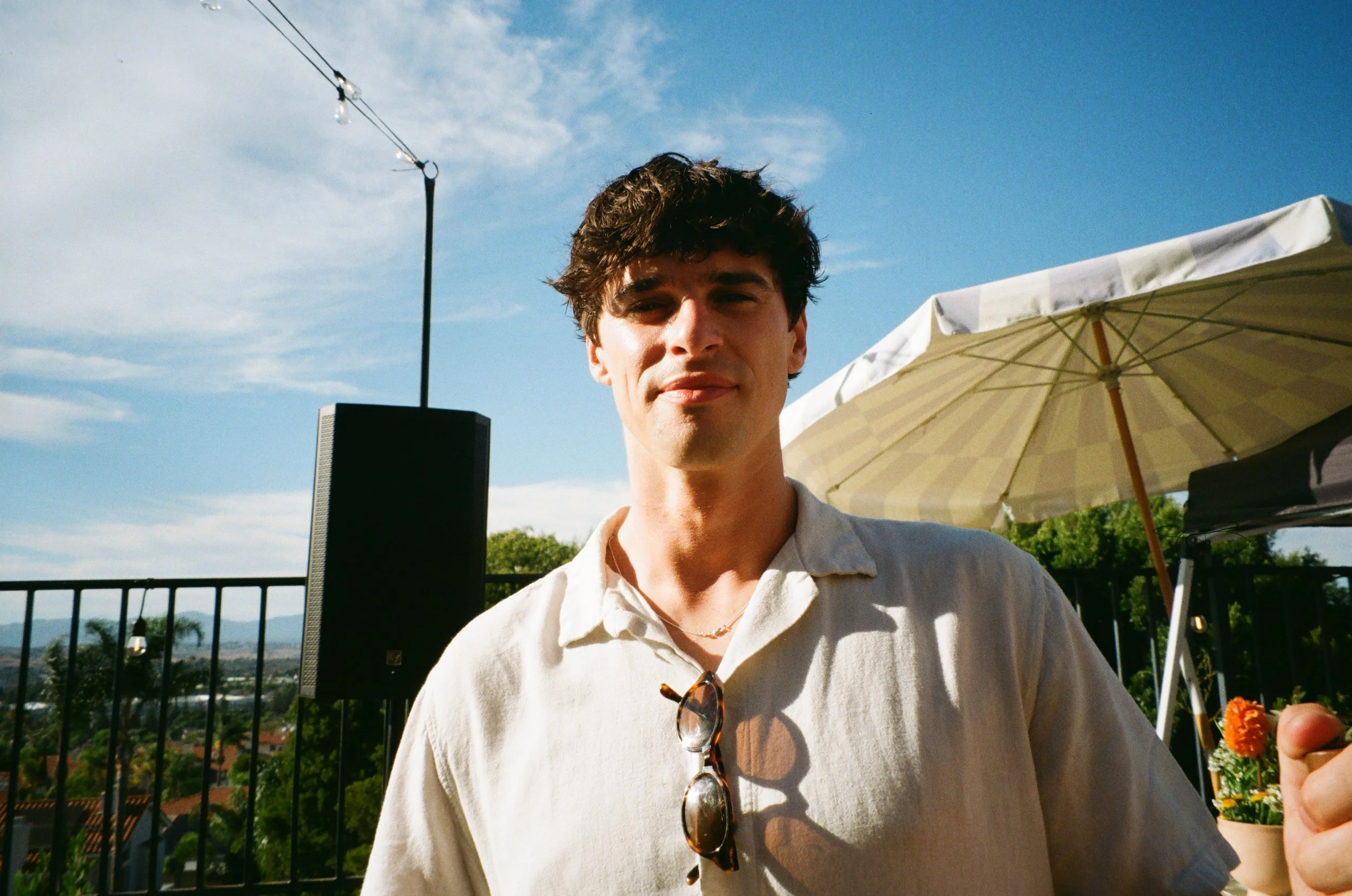 A young man with dark, curly hair and light skin, wearing a cream-colored shirt, standing outdoors on a sunny day with a blue sky, some clouds, a patio umbrella, and a black speaker in the background.