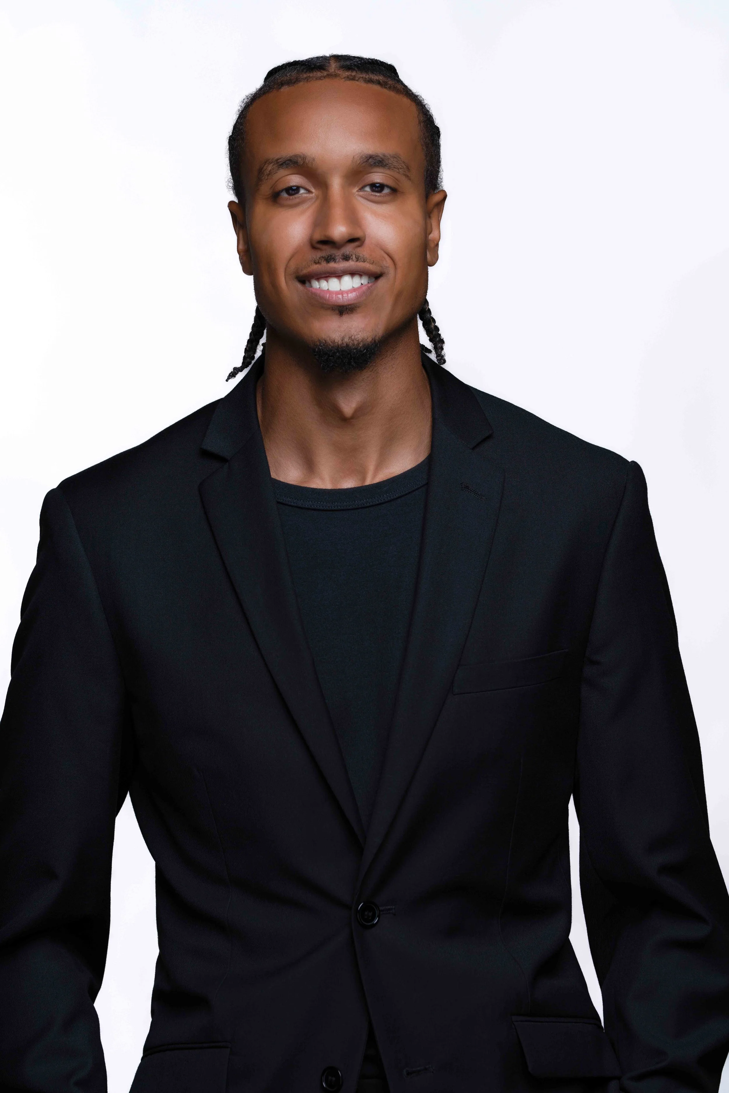 A young man with long braided hair, wearing a black blazer and black shirt, smiling at the camera against a plain white background.