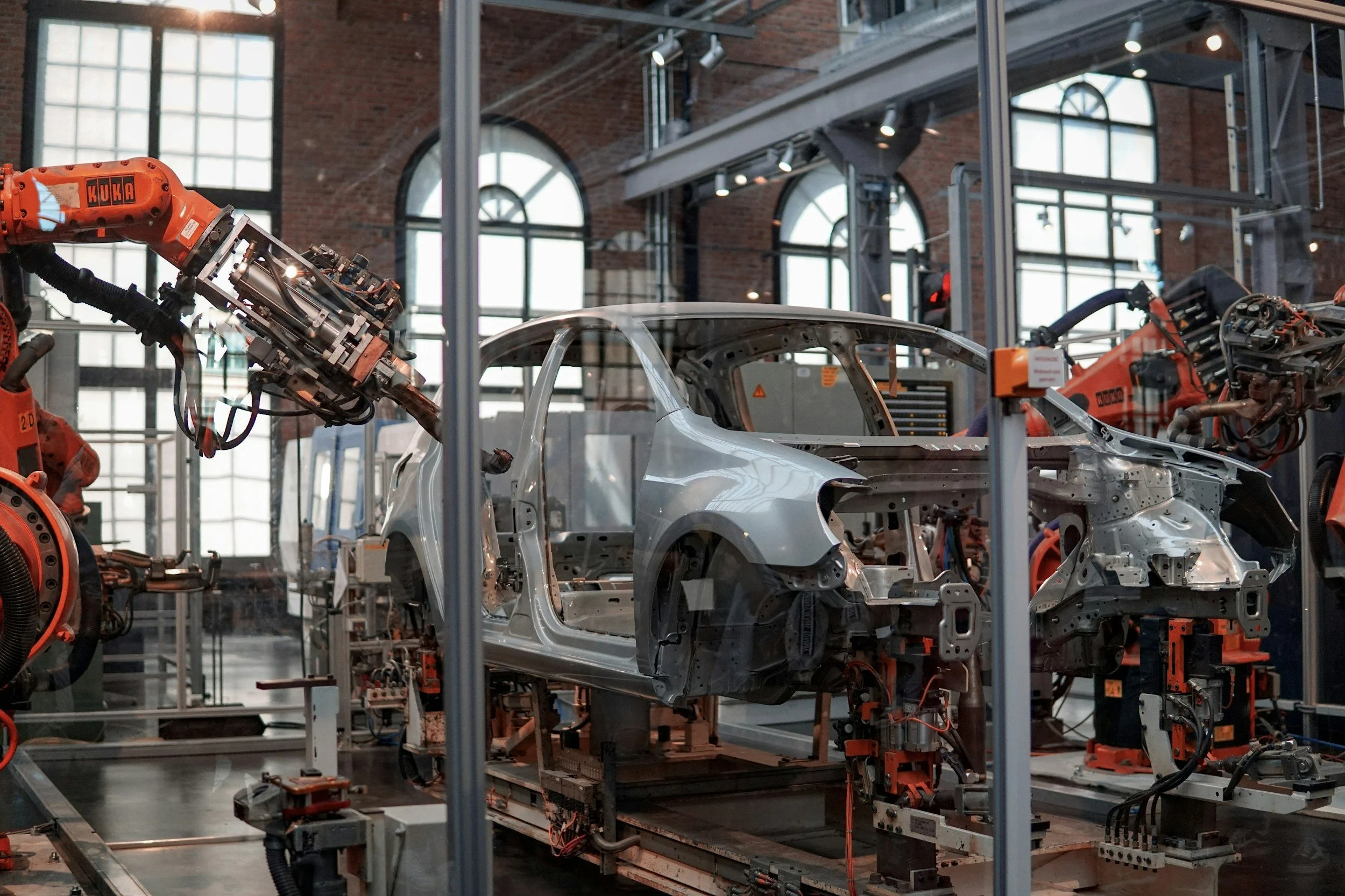 Inside an automotive manufacturing plant, robotic arms assemble a car's body frame within a factory with large windows and brick walls.