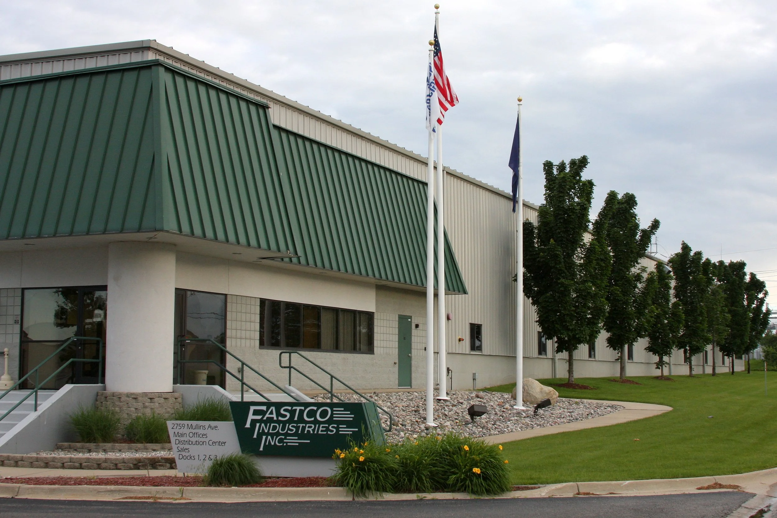 Exterior of Fastco's Sales and Distribution building, three flagpoles with US and company flags, a landscaped area with trees, and a sign reading 'Fastco Industries Inc.' in front of the entrance.