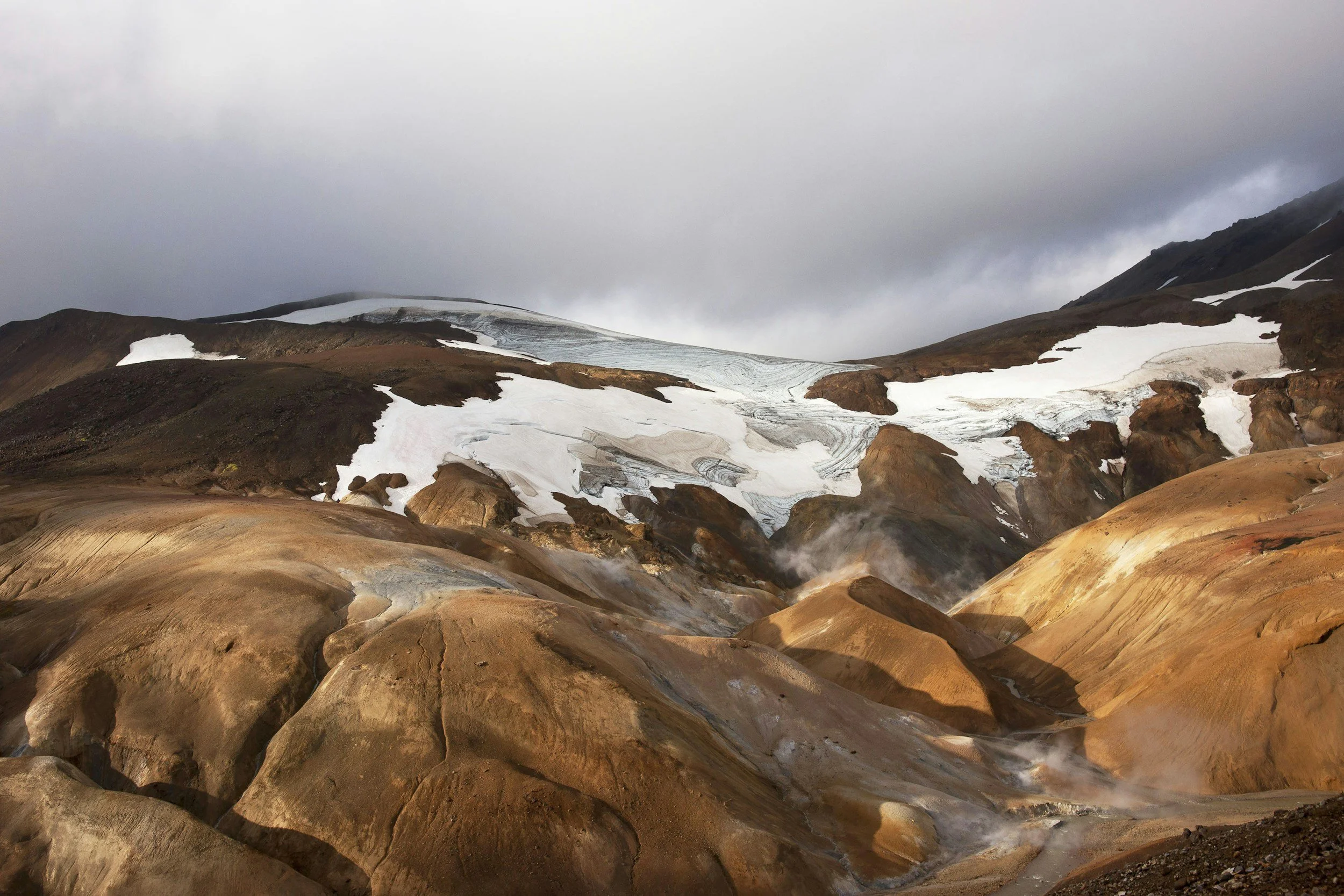 A rugged mountain landscape with snow patches, steaming geothermal area, and colorful rocks under a cloudy sky.