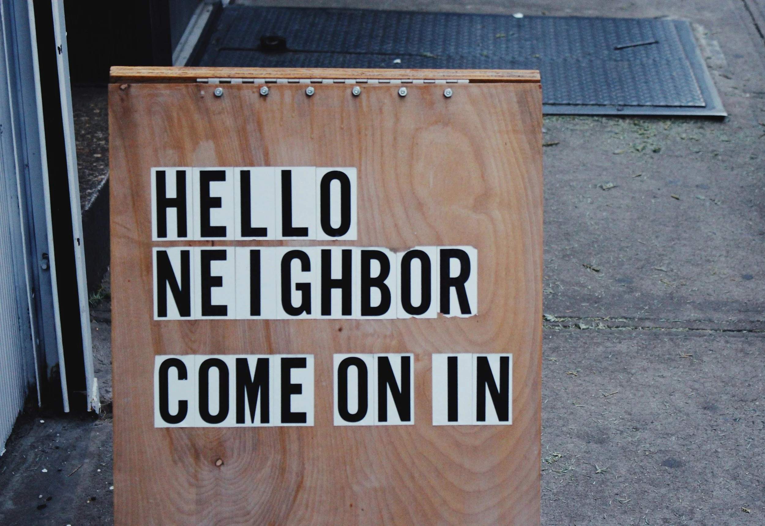 A wooden signboard with the words 'HELLO NEIGHBOR' and 'COME ON IN' written on it, placed on a sidewalk.