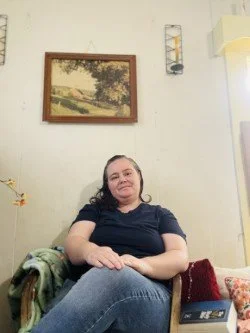 A woman sitting on a chair in a living room with a painting of trees and a path on the wall behind her, holding her hands in her lap and smiling.