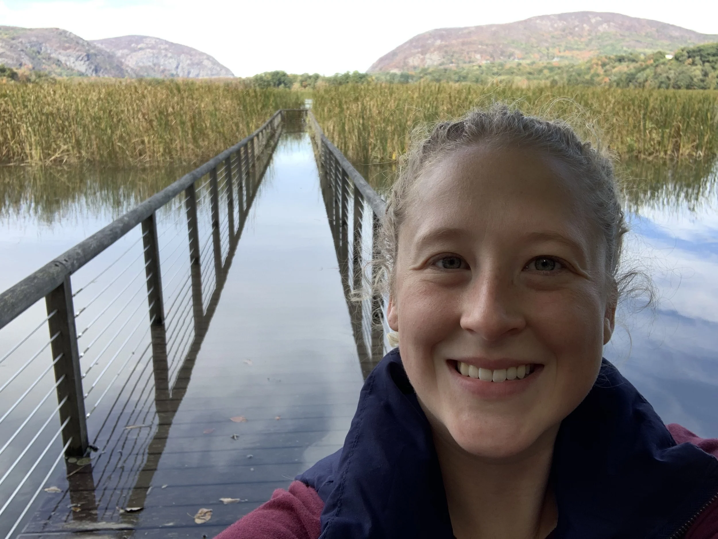 A smiling woman taking a selfie on a wooden pier over a lake with tall reeds, mountains in the background, and cloudy sky.