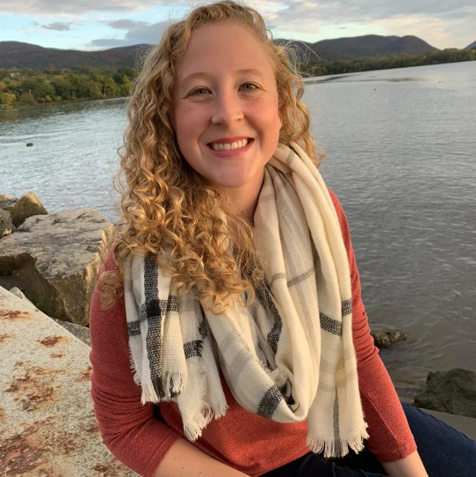 A smiling woman with curly blonde hair sitting by a river with mountain scenery in the background, wearing a red top and a plaid scarf.