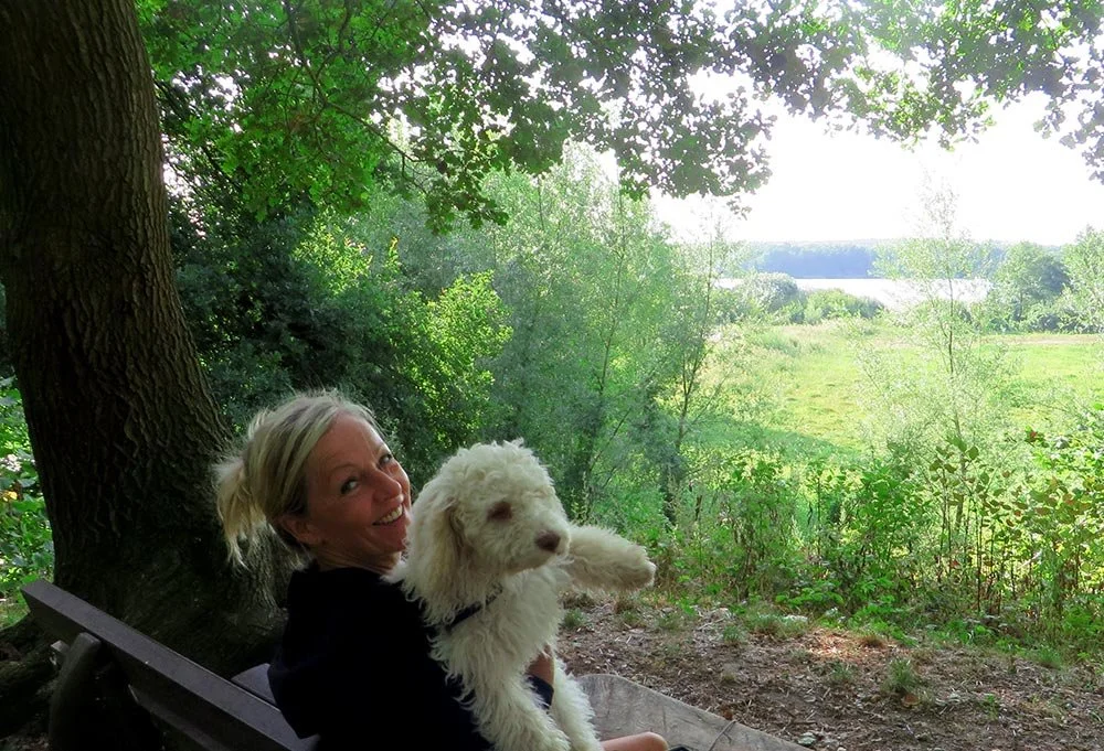 Eine Frau sitzt auf einer Bank unter einem Baum im Grünen und hält einen weißen Hund in den Armen, während sie in die Kamera lächelt.