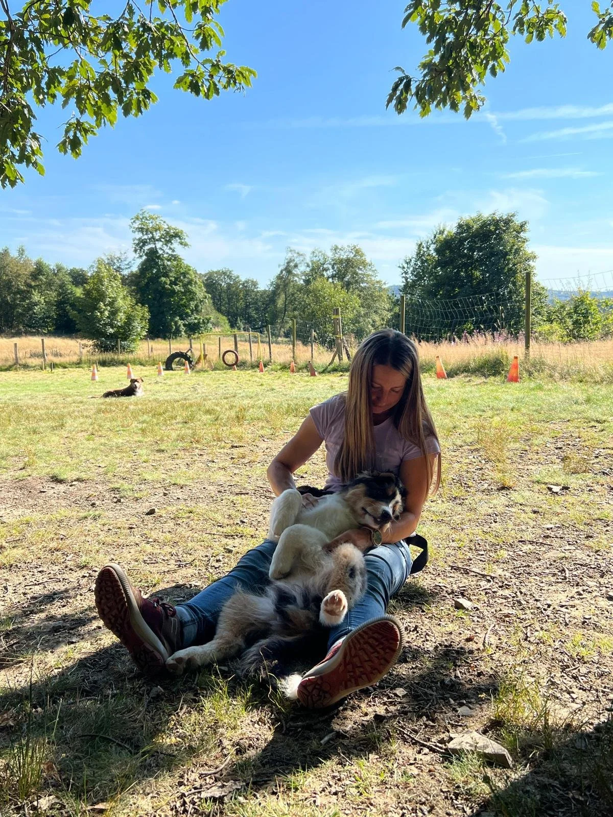 Frau sitzt auf dem Boden im Freien mit einem Hund, der auf ihrem Schoß liegt, während in der Hintergrund ein weiterer Hund auf dem Gras liegt. Es ist ein sonniger Tag, die Umgebung ist grün und es gibt einen Zaun im Hintergrund.