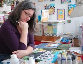 Photo of Jen Niles at her art table with paintings on the wall and on the table.
