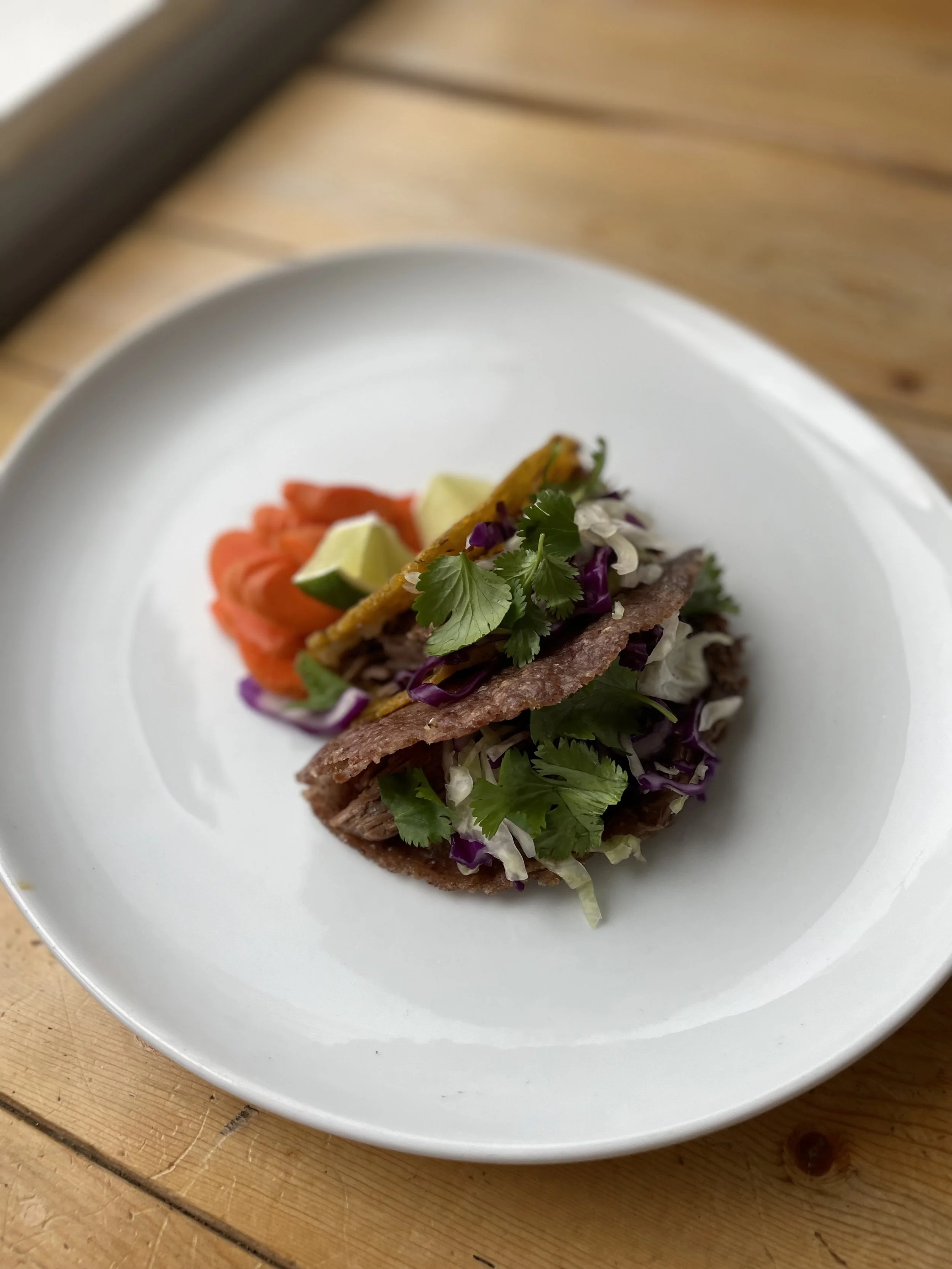 Close-up of two tacos filled with beef, shredded cabbage, cilantro, and red onion, served with lime wedges and sliced tomatoes on a white plate.