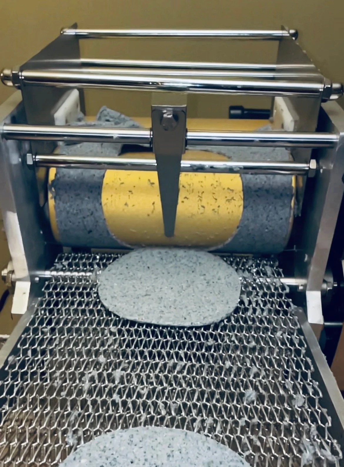 A terrazzo tile making machine with a semicircular terrazzo tile in the foreground and a roller with terrazzo material in the background.