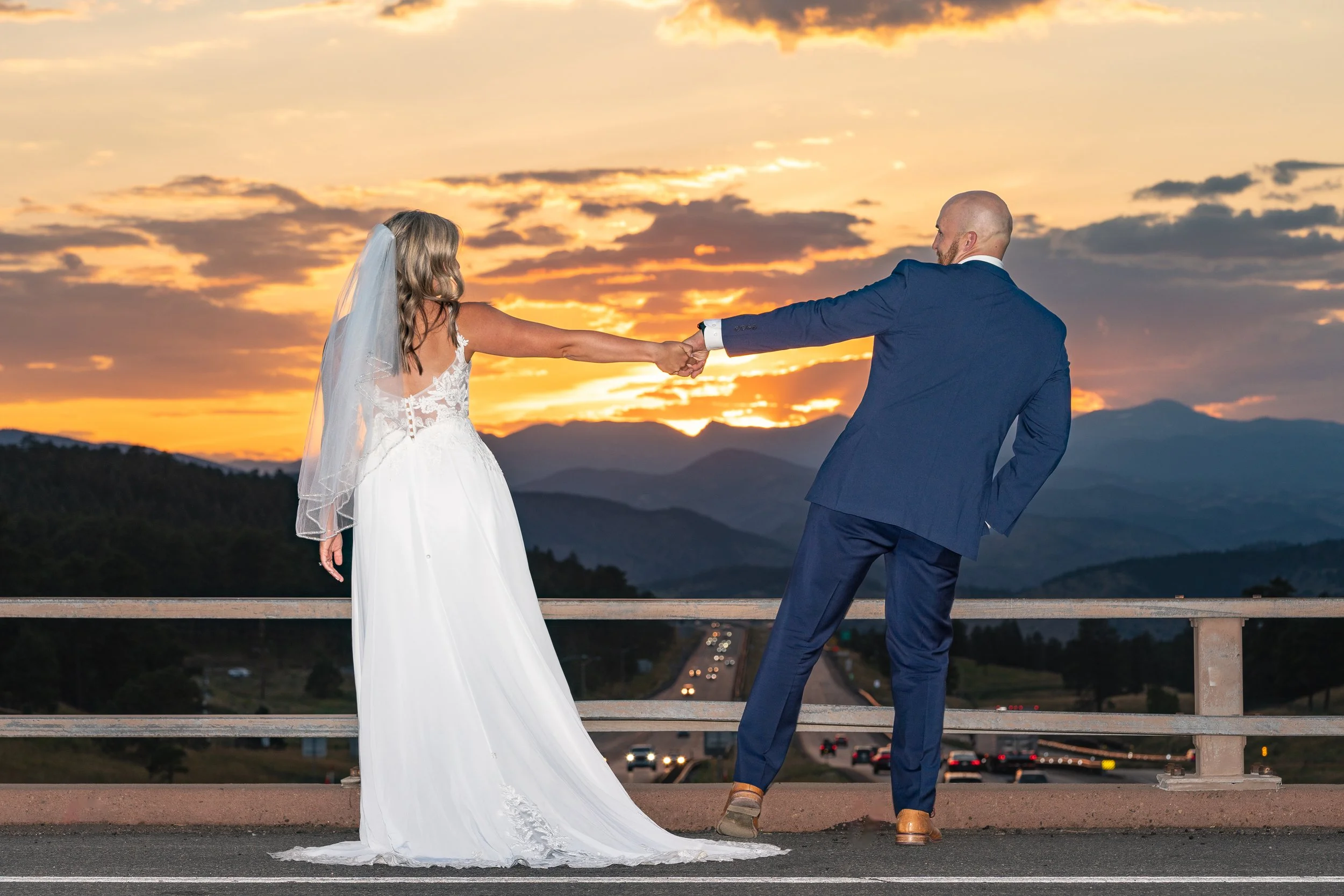 A bride and groom holding hands at sunset on a bridge overlooking a highway and mountain range.