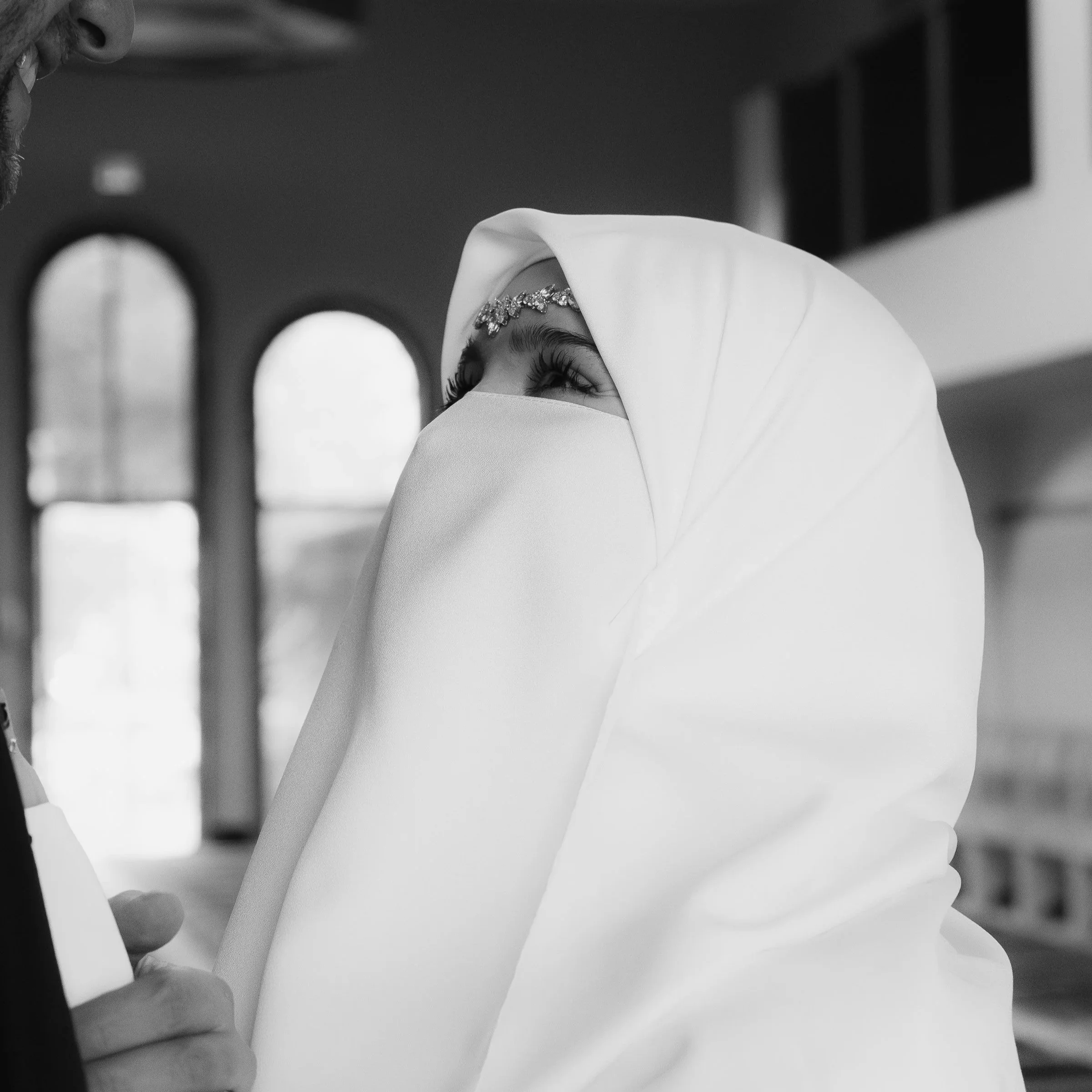 A woman wearing a white niqab and a jeweled headband, looking upwards, inside a church with arched windows.